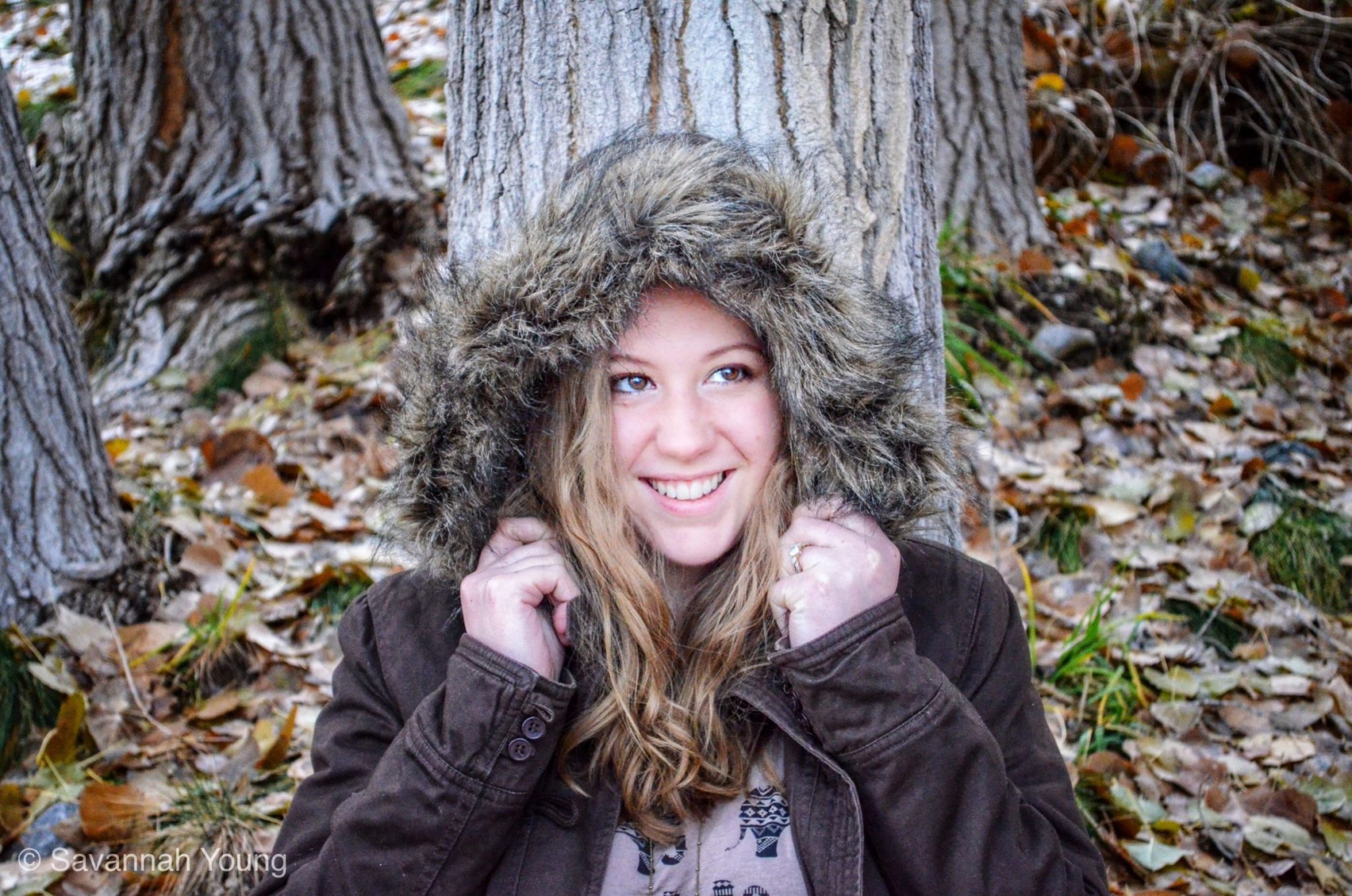 Savvy Leigh Photography. Alison in the forest trees with a fuzzy fur hood on her coat. She is smiling and looking beautiful. Taken in Provo, Utah Wedding Photographer.