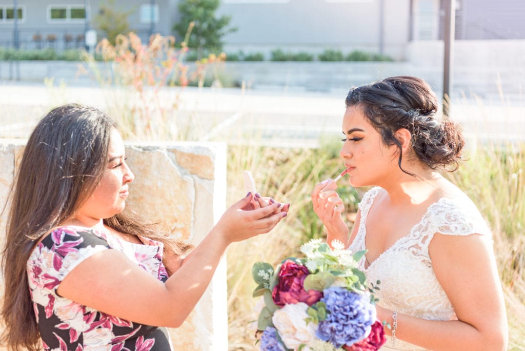 A bride touching up her lipstick that she kept in her wedding day emergency bag. Her maid of honor is helping her.