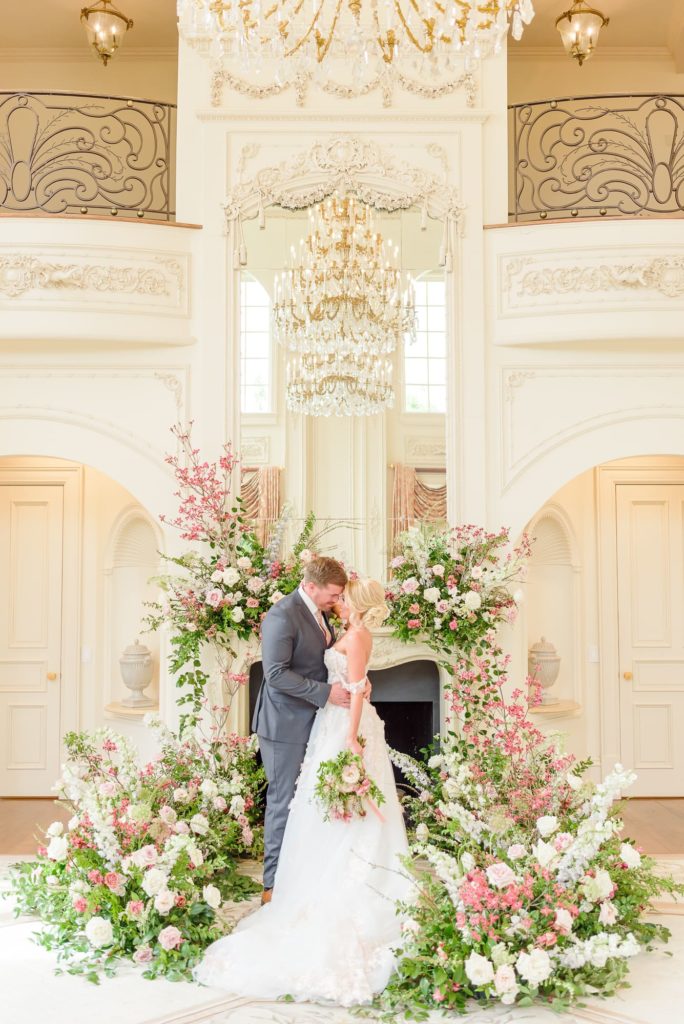 A bride and groom stand in front of a fireplace adorned with flowers at their elegant mansion wedding.