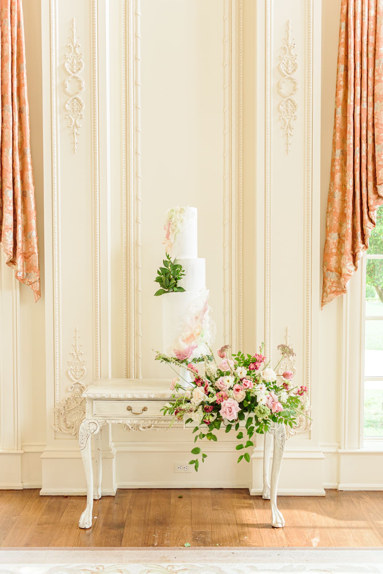 A tall, tiered cake stands on top of a desk overflowing with flowers at this elegant mansion wedding.