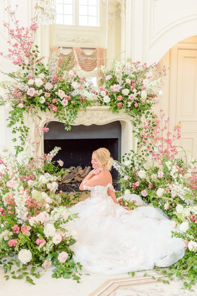 An elaborate floral wedding decor display at a pink and white wedding. Pink and white flowers surround an ornate fireplace as a bride sits in front of it.