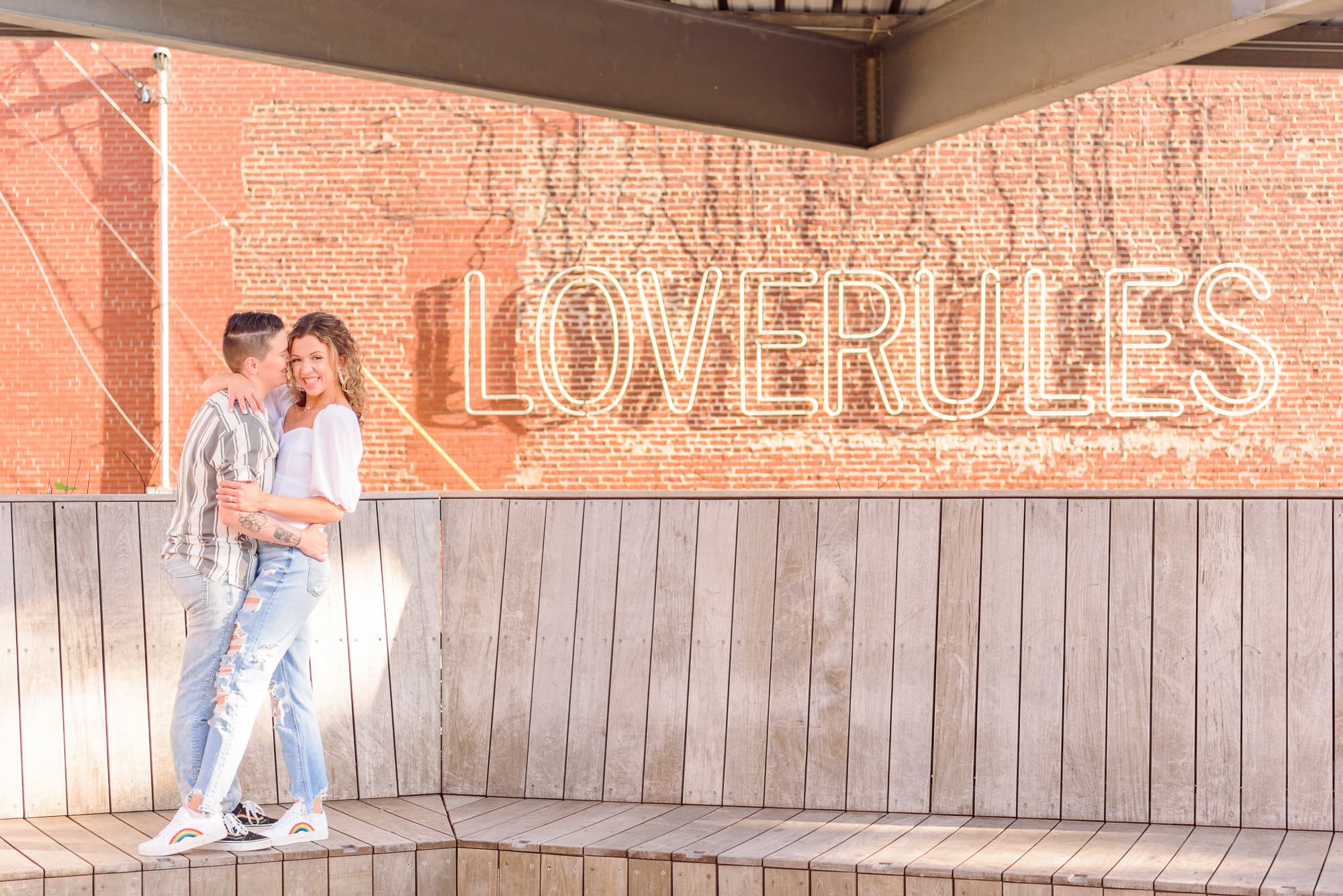 A lesbian couple takes their spring engagement photos in front of a sign that says "Love Rules".