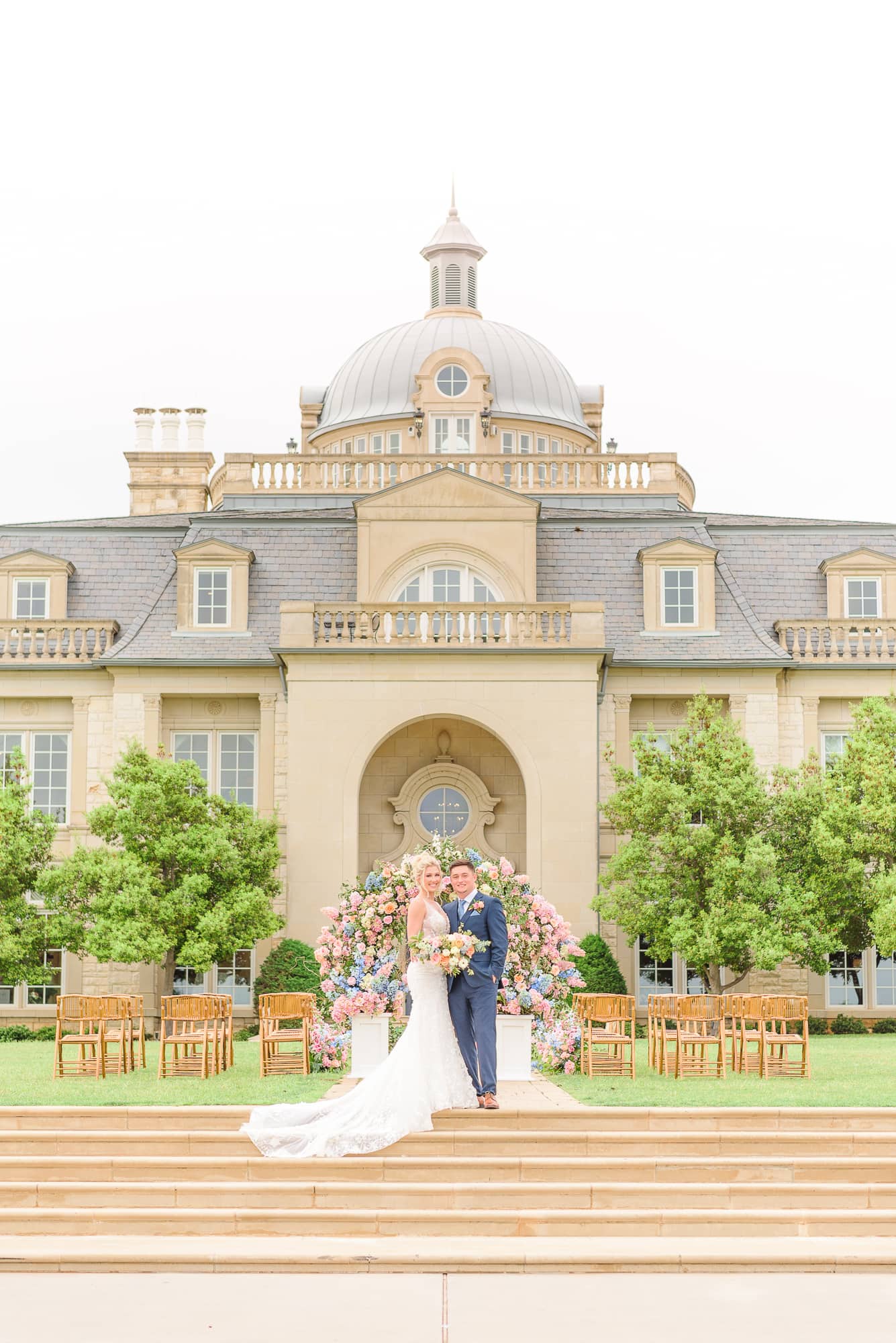 The bride and groom are surrounded by summer colors as they stand in front of their wedding venue.