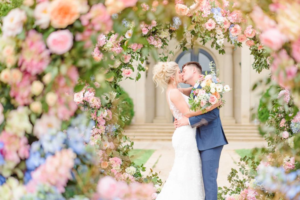 A first kiss picture during the wedding ceremony where the couple is surrounded by a flower arch.