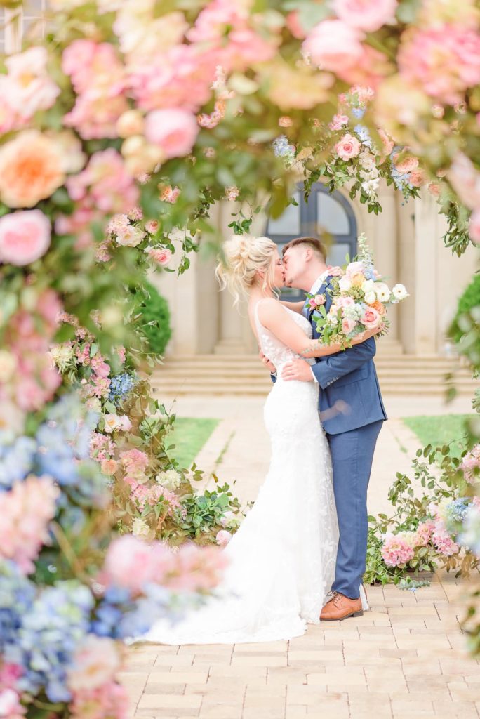 Floral wedding decor can be used to create ceremony arches, like this couple sharing their first kiss underneath pink and blue blooms.
