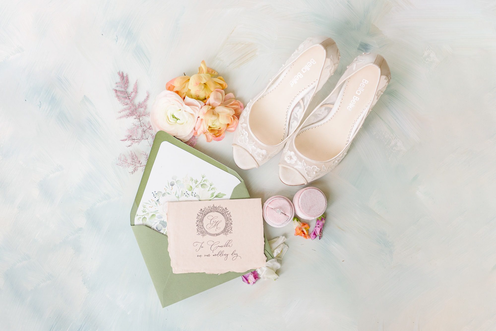 A green envelope, purple flowers, and white wedding shoes are styled artistically.