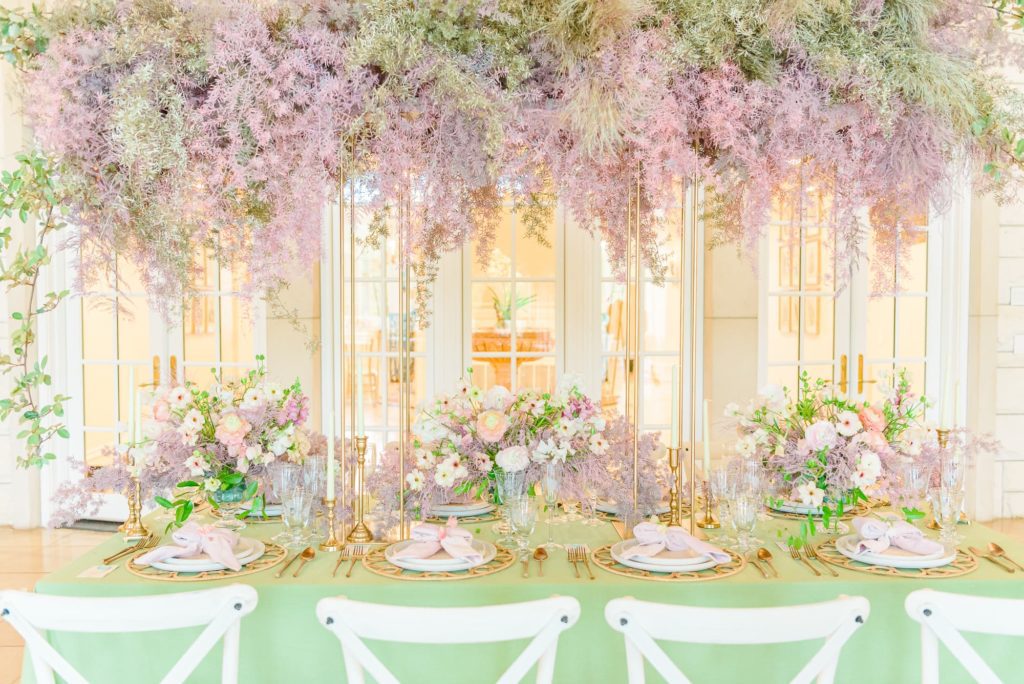 A lavish table setting looks to be exploding with purple flowers, and floral wedding decor is raised up on gold stands so the flowers are suspended above the table.