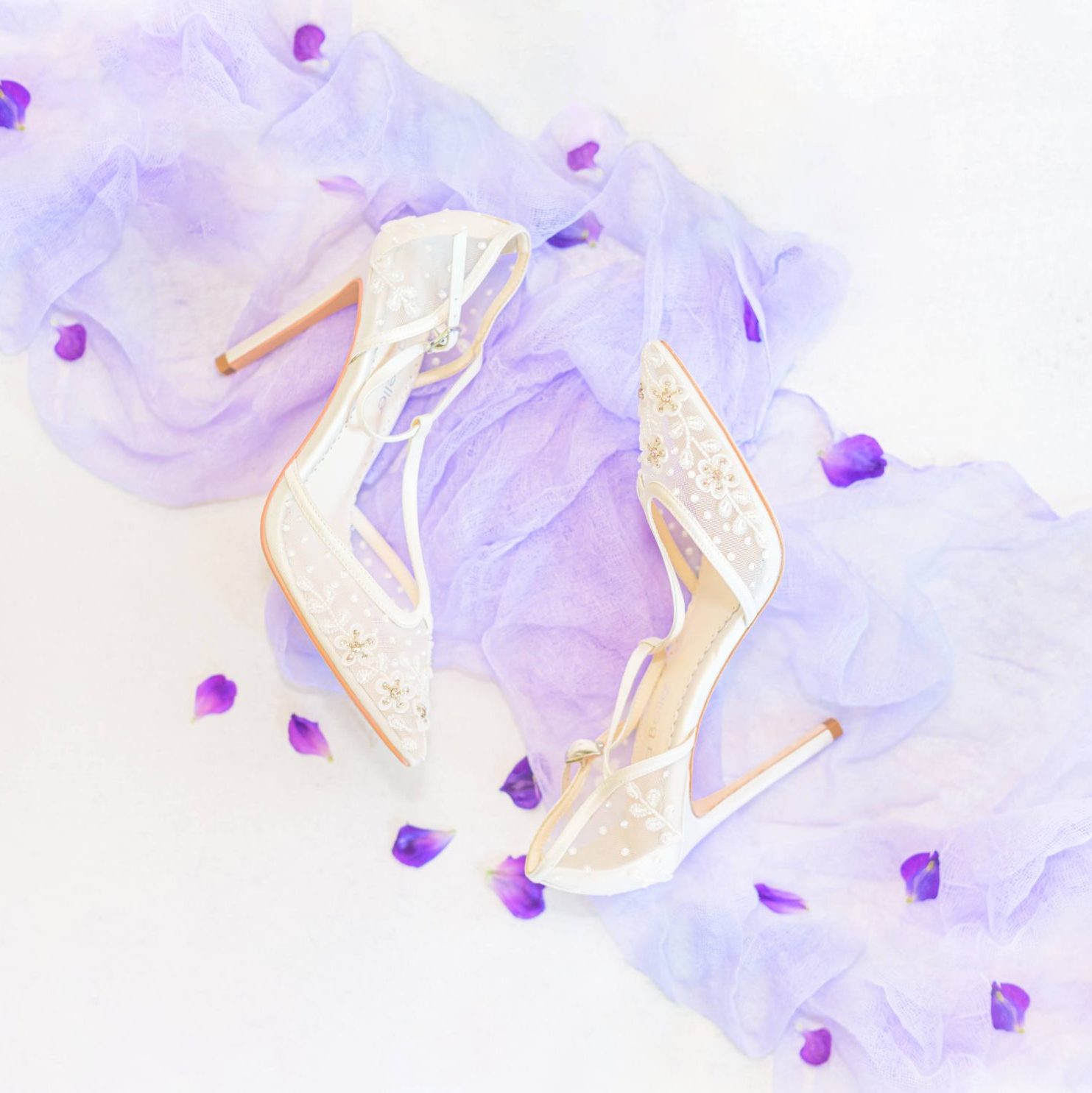 Wedding shoes lying on top of purple ribbon with flower petals scattered around.
