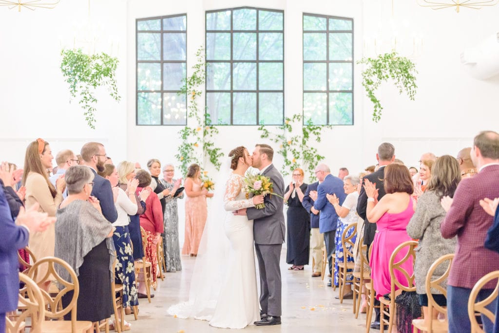 Cecilia and Ethan kiss at the end of the aisle while their friends and family applaud in the background of their first kiss picture.