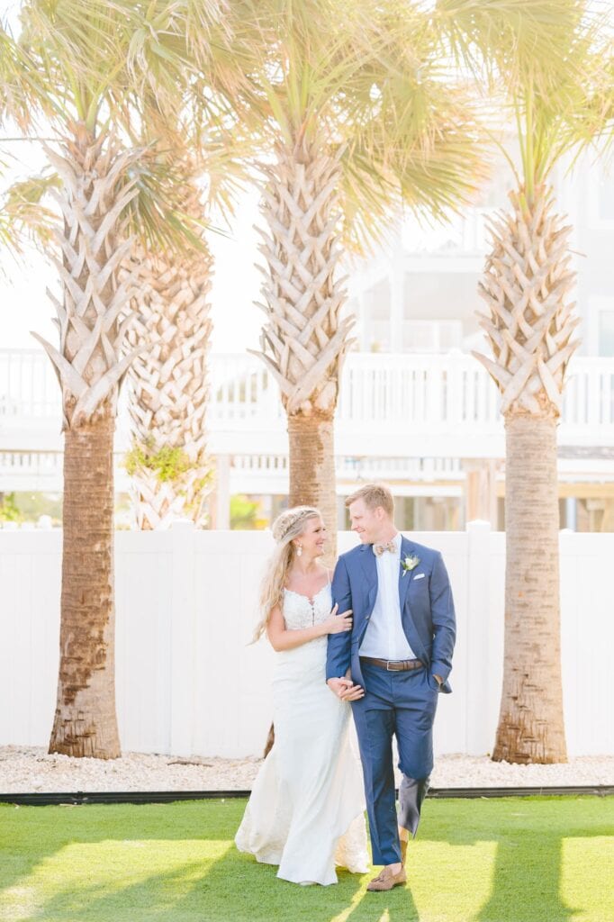 The bride and groom walk away from a line of palm trees during their wedding at the beach in North Carolina.