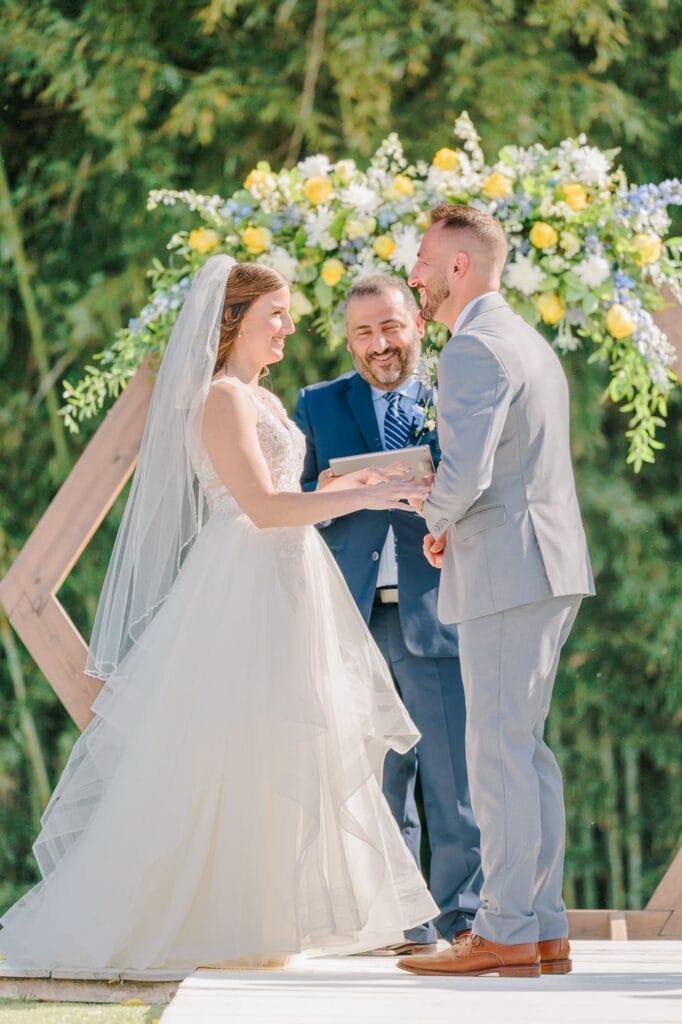 The bride and groom give their vows outside in front of an arch adorned with flowers at their outdoor North Carolina wedding.