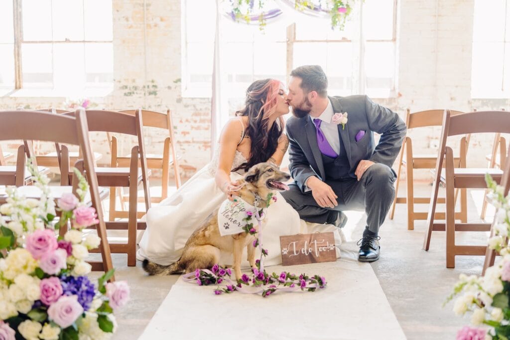 The bride and groom kiss in front of their ceremony arch at the Kettle Room and hold their dog in front of them.