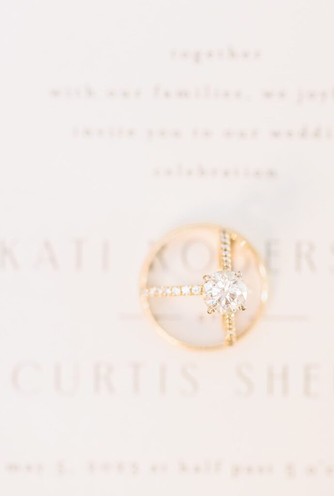 Beautiful gold engagement rings sit on a wedding invitation inviting guests to a celebration at the Ruth in Charlotte, NC.