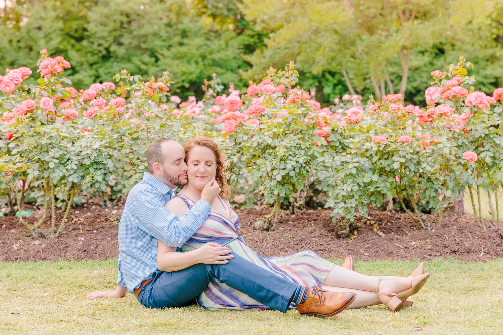 The couple sits on the ground and holds each other in front of the blossoming roses at the rose garden in Raleigh, NC.