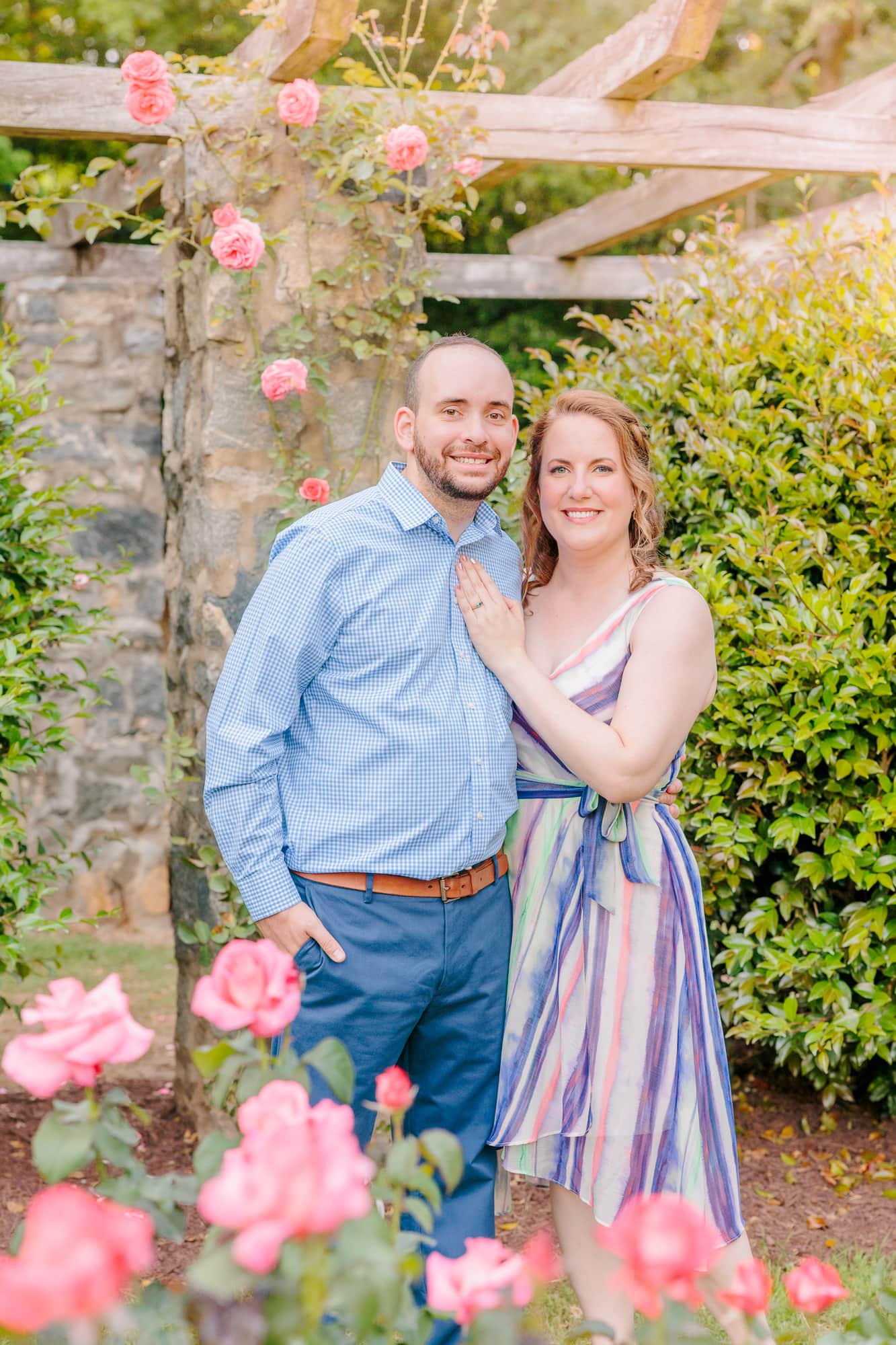 The couple smiles for the camera, posing in front of a stone pillar at the Raleigh, NC rose garden.