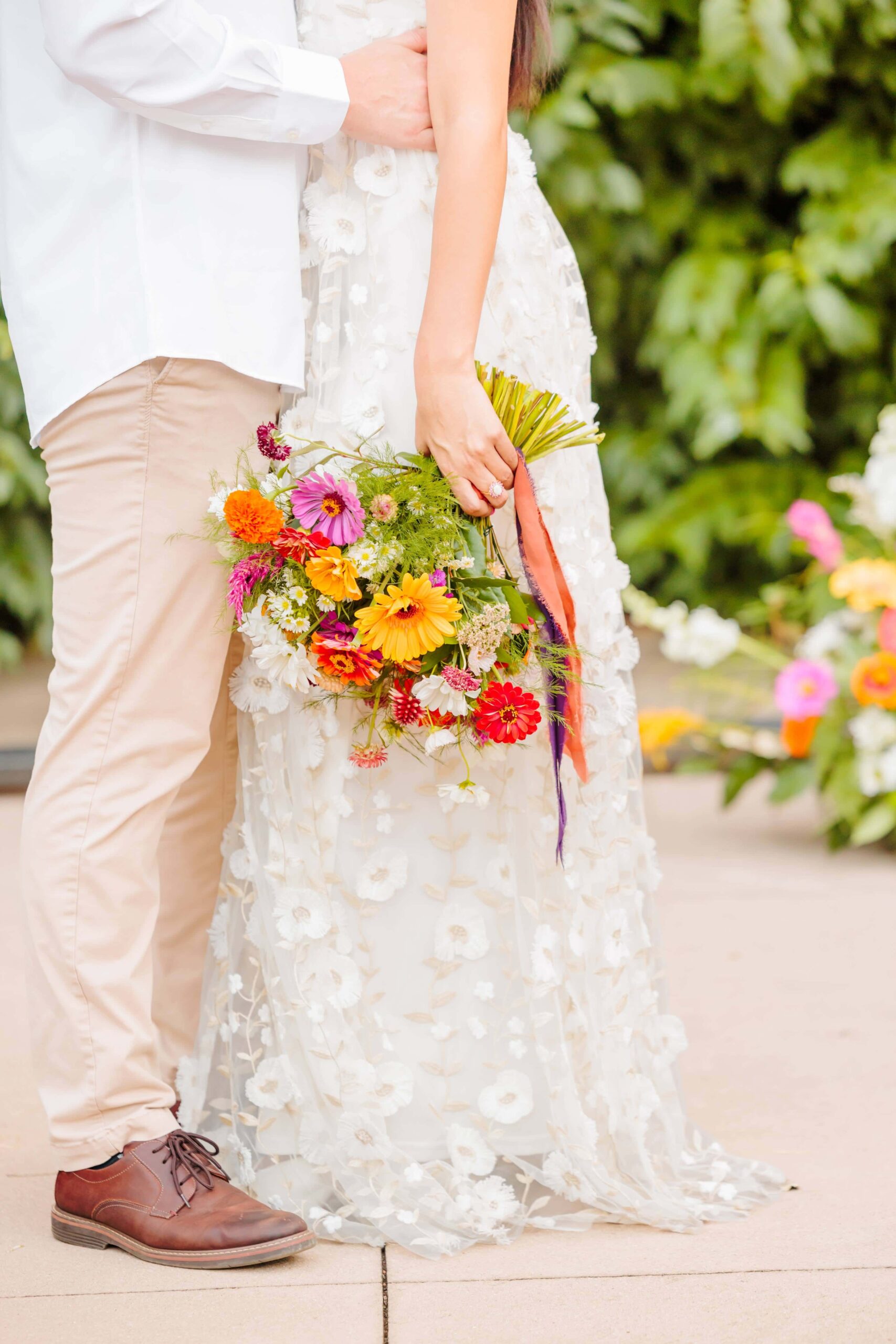 The bride holds her bouquet down by her side and you can see embroidered flowers on her disco wedding dress.