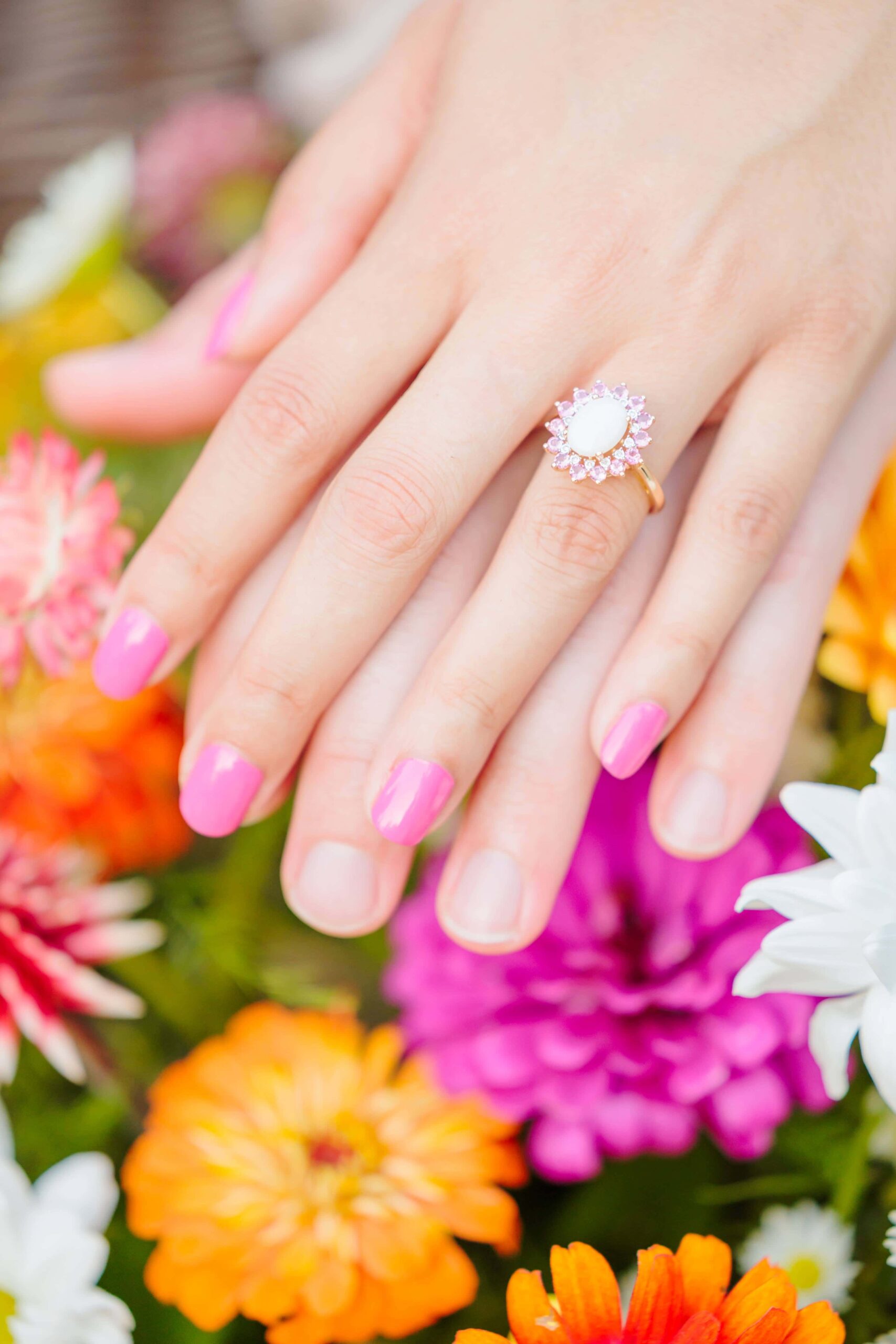 A disco wedding ring with an opal surrounding by pink stones is shown with bright flowers in the background.