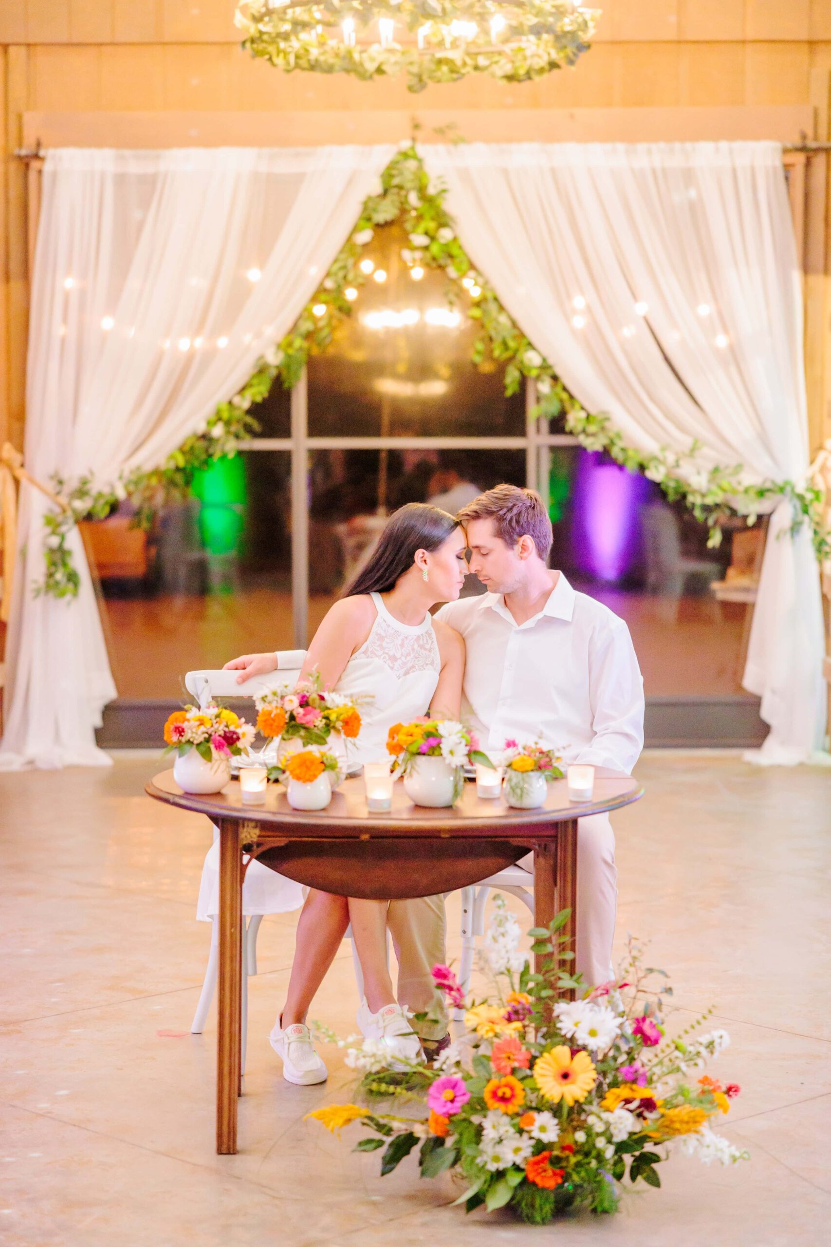 The bride and groom sit down at a disco themed sweetheart table surrounded by florals at their wedding.