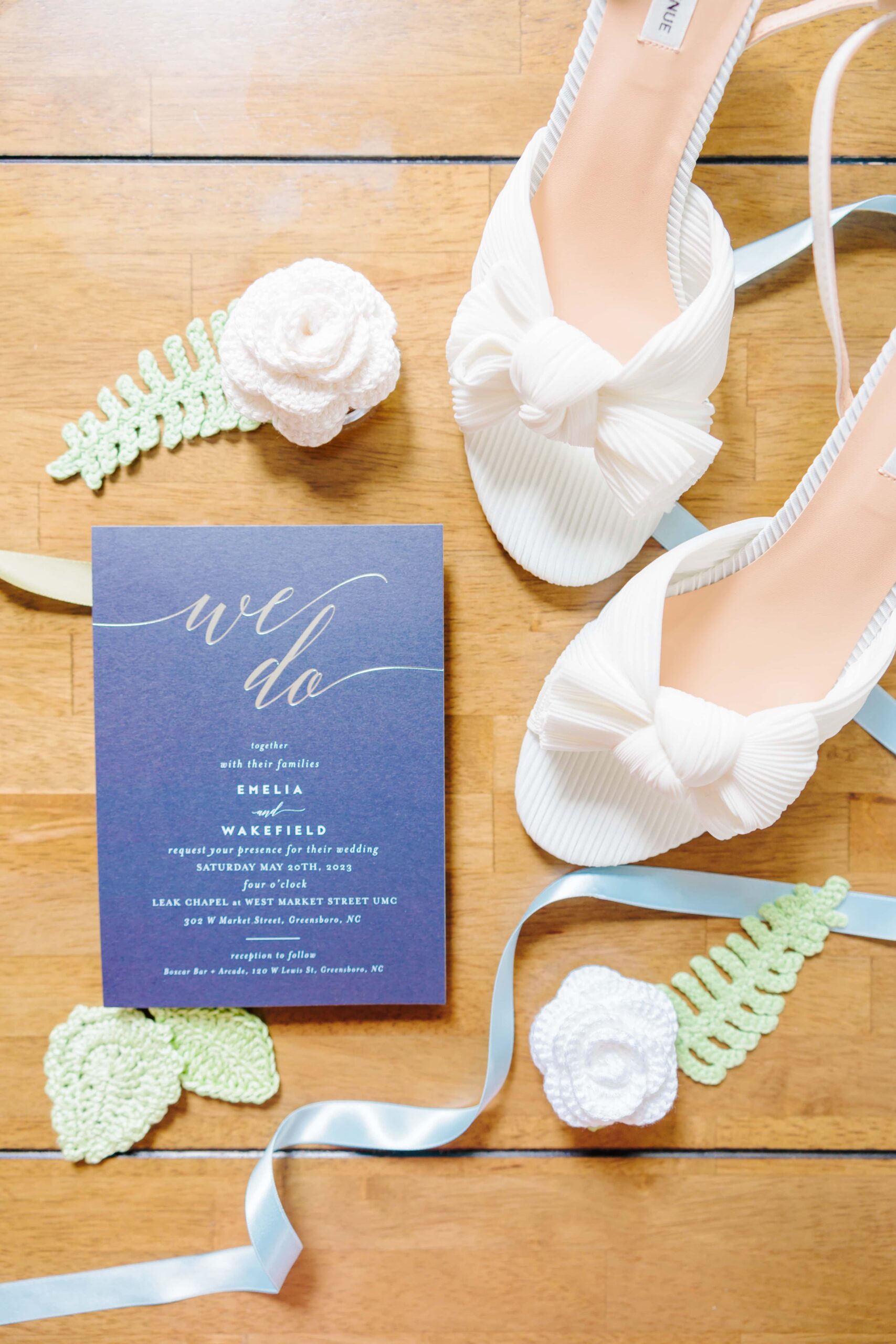 A dark blue wedding invitation inviting guests to this Greensboro wedding is placed amongst crocheted flowers, blue ribbon, and the bride's shoes.