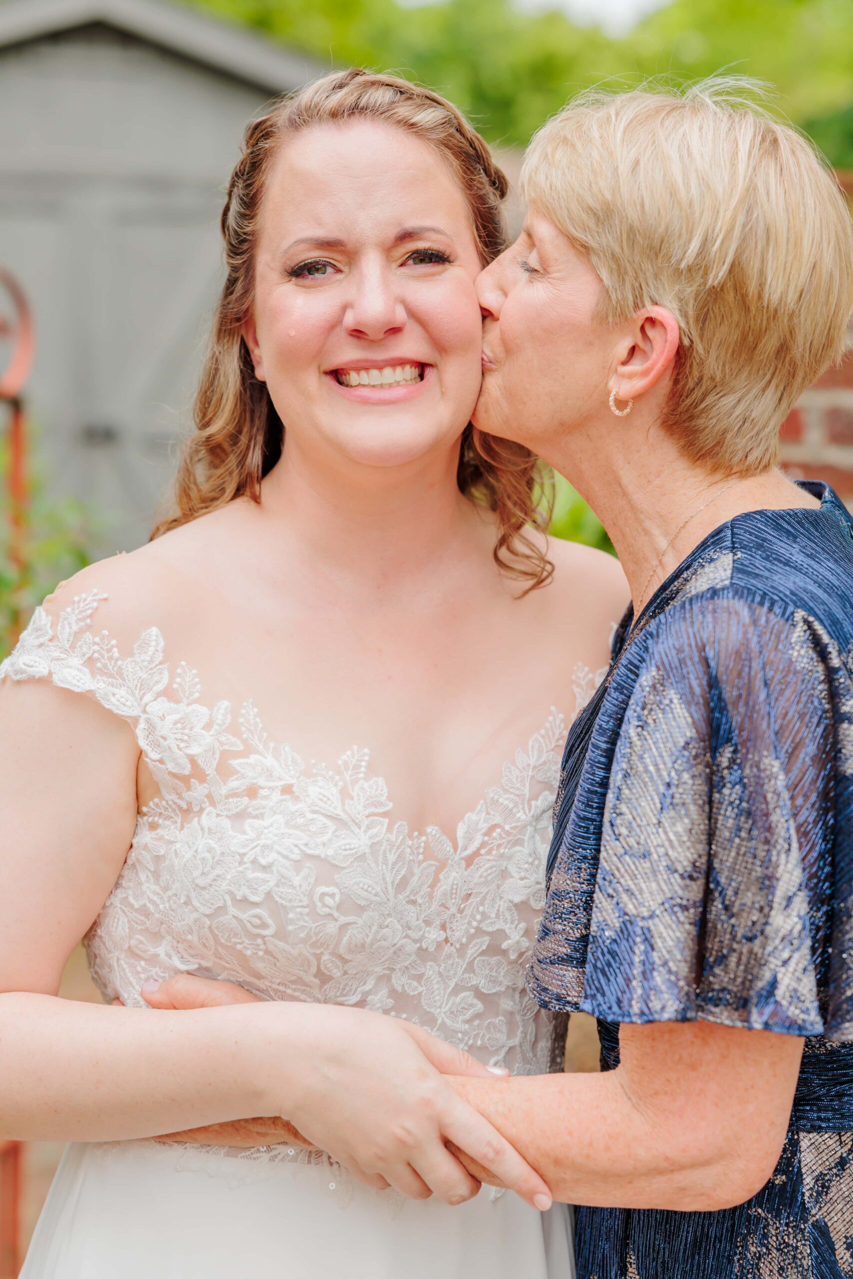 The bride smiles to the camera with tears in her eyes as her mother kisses her cheek before her wedding day in Greensboro.