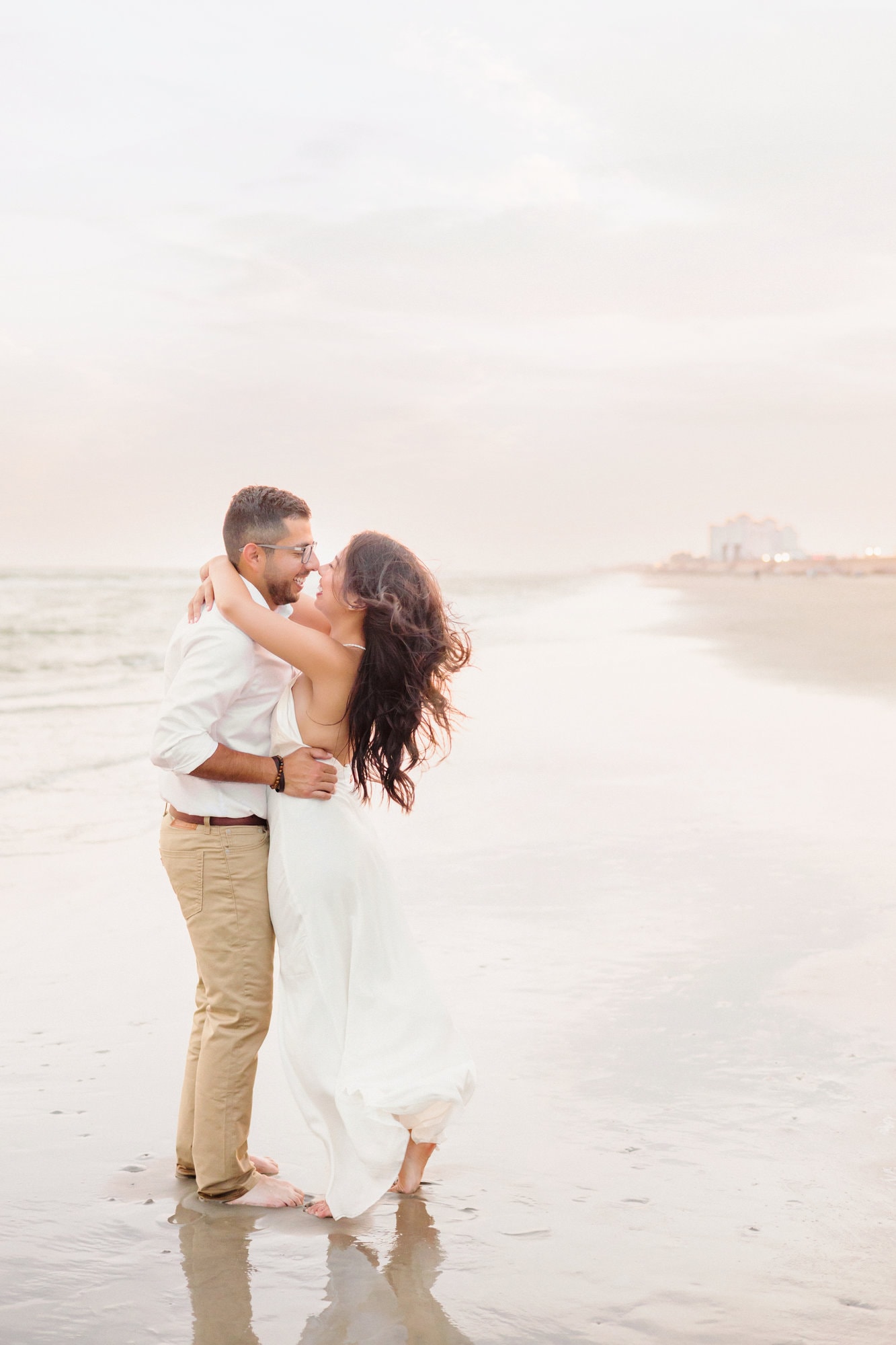 The couple twirls on the sand during their engagement pictures on the beach as the wind whips her hair and dress.