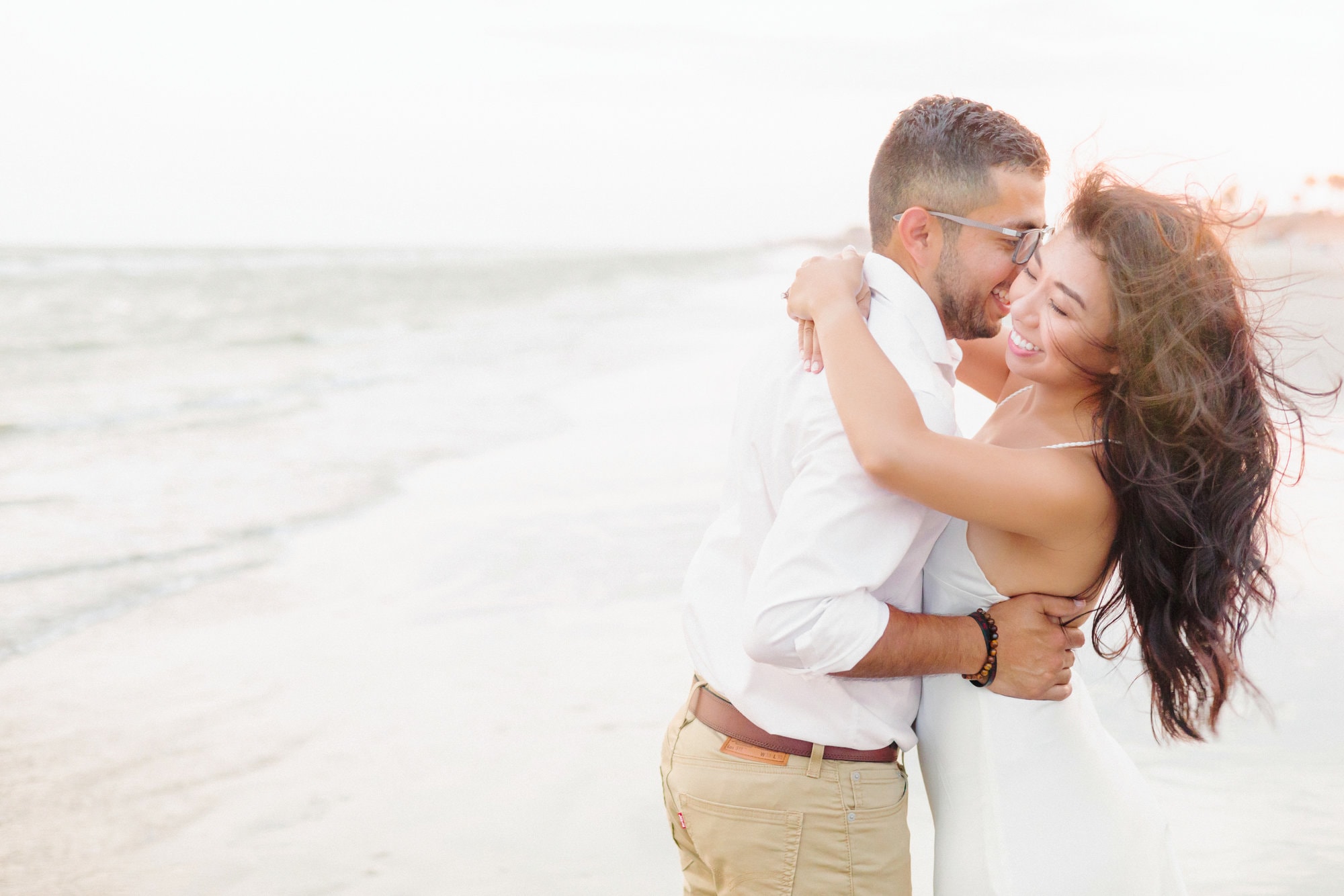 The couple holds each other close and giggles for their engagement pictures, while the calm waves of the beach are shown in the background.