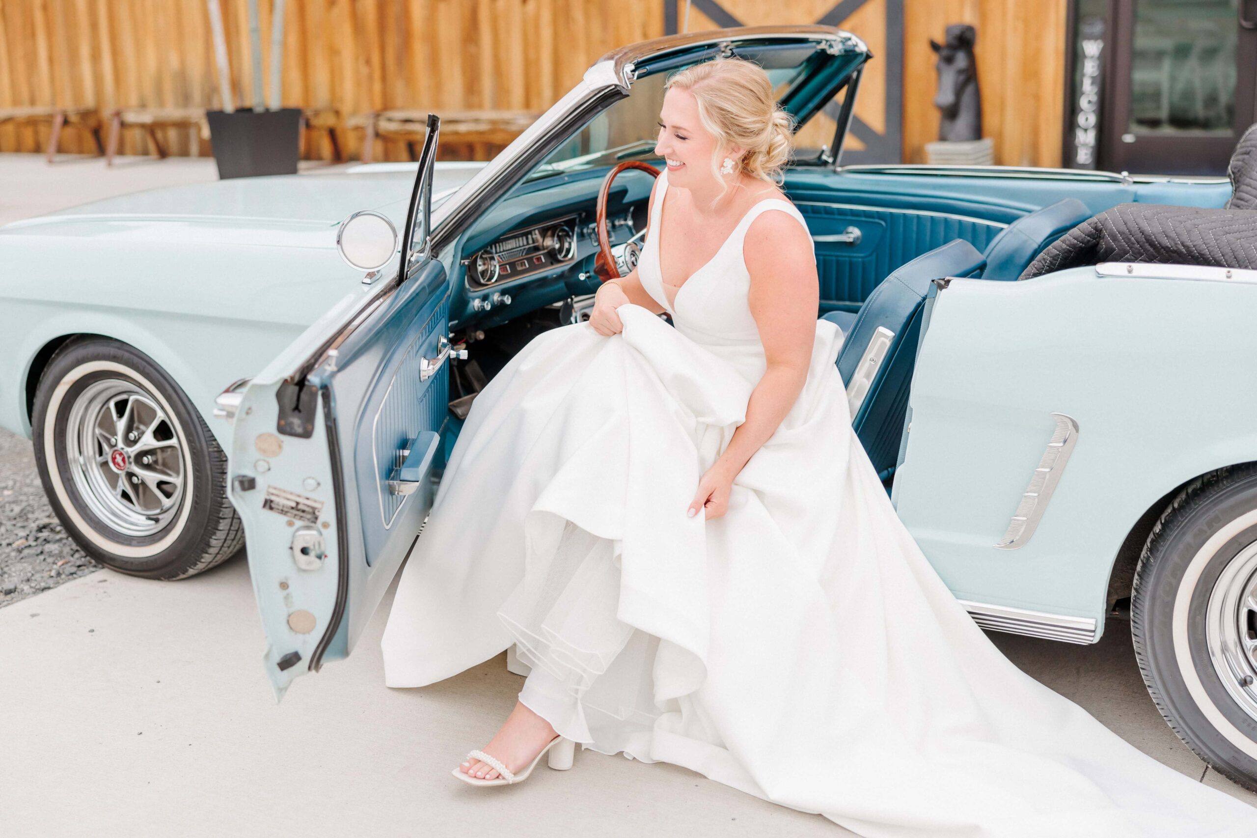 A bride holds her dress as she exits a classic blue car.