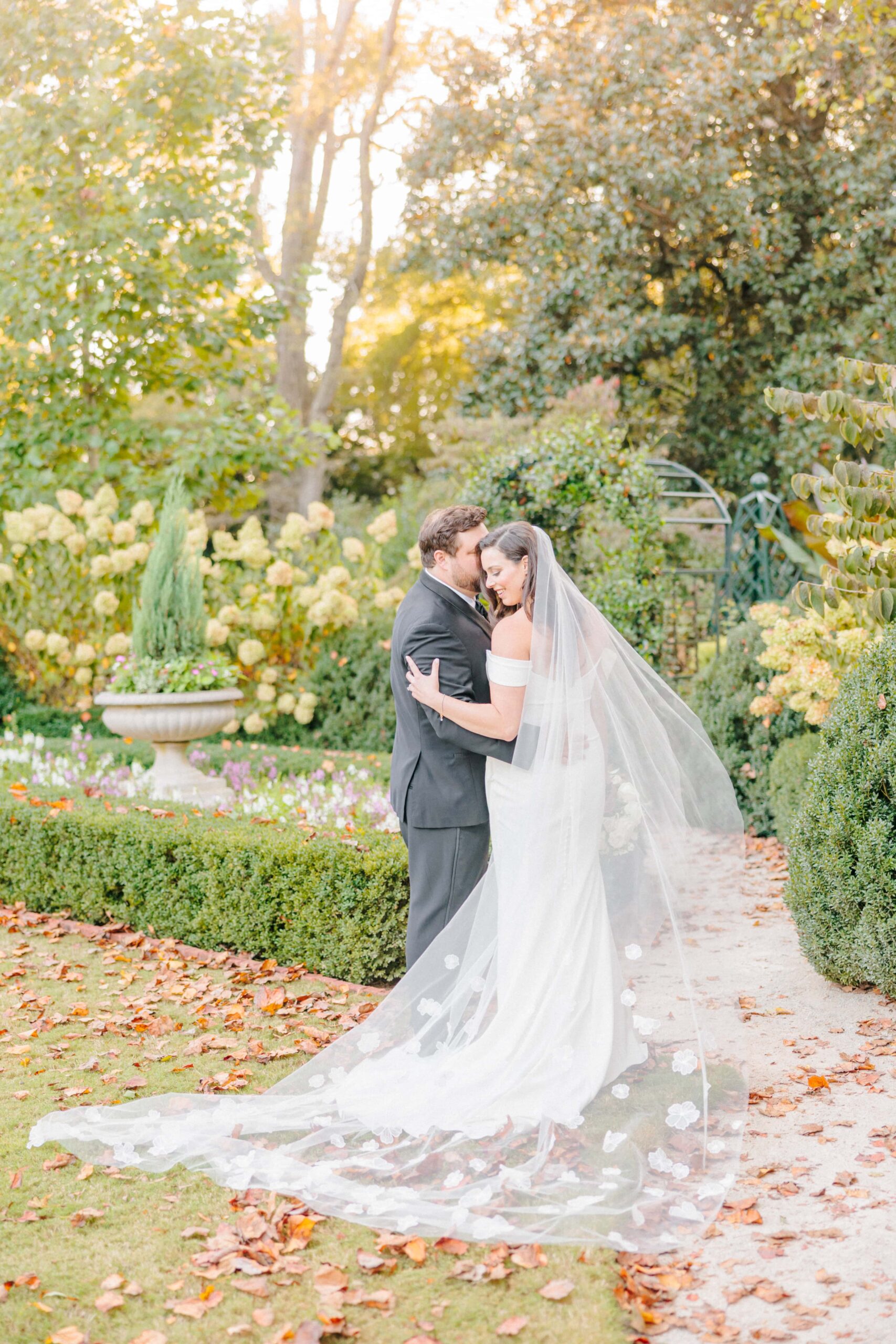 The groom holds the bride tenderly for their wedding photos, and her textured veil blows on the lawn of the Duke Mansion.